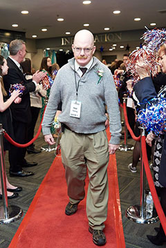 person walking toward the camera. They have no hair and are wearing glasses, a grey sweater and dark pants. They are walking on a red carpet with people clapping on either side.